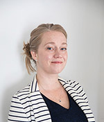 Three-quarter-view headshot of Shana MacDonald, a woman with long blond hair tied back in a loose bun, wearing a black-and-white striped jacket and slight smile